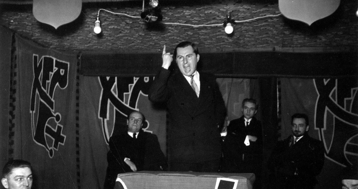 Léon Degrelle en discours, le doigt levé, devant des bannières Rex